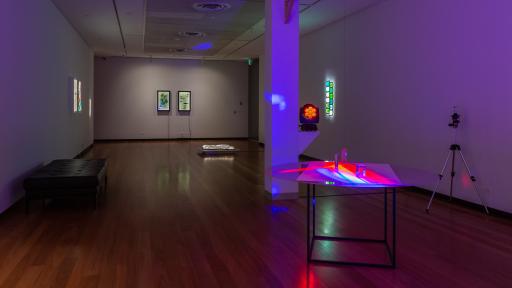 Long thin rectangular art installation space with a square column and hardwood floors. There is a round-top table with light-reflecting objects which create a purple and orange pattern. There is a tripod to the right.