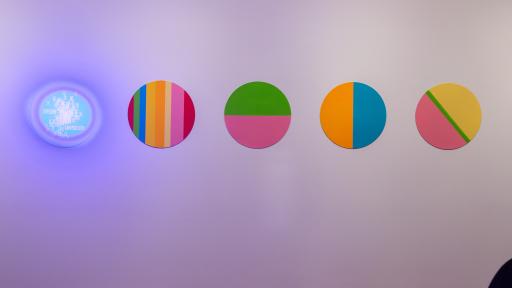 Art installation featuring five circles containing straight lines and a mix of pink, orange, blue, green and yellow. The circle to the left is blue with white words and X symbols.
