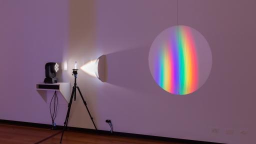 Art installation featuring a projector and tripod pointed at a wall to create a round shape with rainbow-style colours
