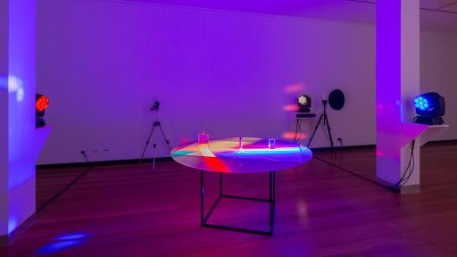 Art installation featuring three light projectors pointed at a round-top table, creating a mix of red, blue and yellow. There is a tripod near the far wall.
