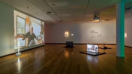 Large indoor art installation space with hardwood floors and three scattered televisions. To the left there is a photo of an elderly woman with her arms outstretched.