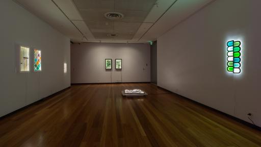 Long thin rectangular art installation space with hardwood floors. There is a work with blue, green and white semi-oval shapes to the right. There are two rectangular works on the left and far wall.