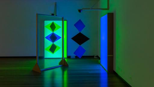 Art installation featuring three diamond shapes hanging from a fitting, and three diamond shapes on a glowing light green background.
