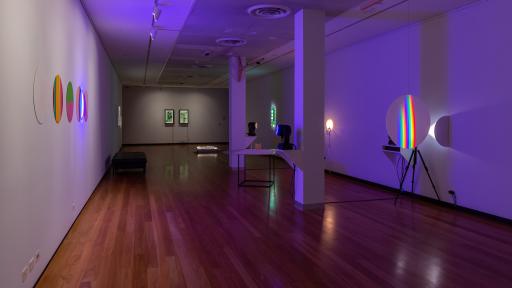 Long thin rectangular art installation space with two square columns and hardwood floors. There are multi-coloured artworks on round backgrounds to the left and right and two rectangular works in the distance.