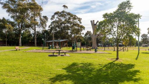 Grass area with playground and set of tables and chairs. There are tall trees in the distance, a small tree in the foreground and a large grey tree stump next to the playground.