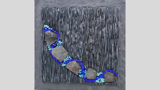 An artwork featuring a mosaic of natural grey stones vertically arranged close together, with a row of 5 larger rounded stones diagonnally across the picee with bright blue and light blue pieces curving their way between the stones