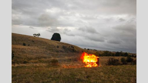Field of dry grass with a piano engulfed in flames towards the right. There is a hill to the far left and a single large tree in the distance.