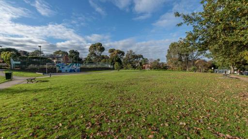 Large grass area with fallen leaves, with trees to the right and in the distance. There is a painted mural, a park bench and a footpath to the left. 