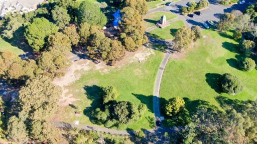 Aerial view of grass area with scattered trees and footpaths in three directions. There is a playground, concrete memorial structure and carpark at the top.