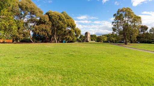 Large grass area with several large trees in the distance. There is a footpath to the right leading to a concrete memorial structure.