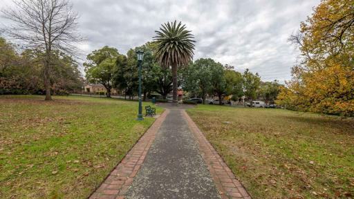 Straight walking path with grass and fallen leaves on either side, leading to a small circular feature with a tall tree. There is a lamp post and park bench to the left of the path.