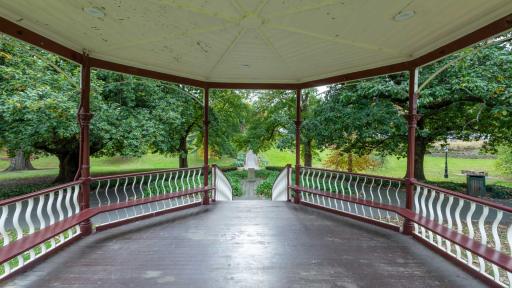Inside view of round outdoor rotunda with steps down to path and large trees. The columns and bench seats are dark red and the roof and balustrades are white.