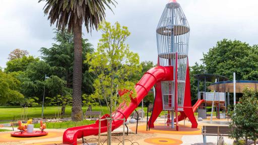 Playground with rocket-shaped climbing feature and slide, and small carousel and park bench. There are several trees in the playing area and the background.