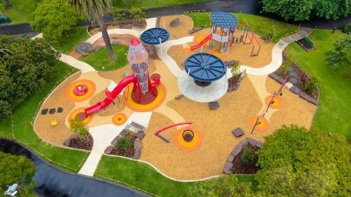 Aerial view of playground with rocket-shaped climbing feature and slide, and three other features with round tops. Grass and several large trees surround the area.