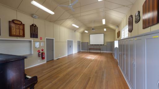 Inside of a hall with polished floorboards and a projector screen at the far end. There are three grey doors along the left and grey cupboards along the right. Part of a piano is visible at the bottom left.