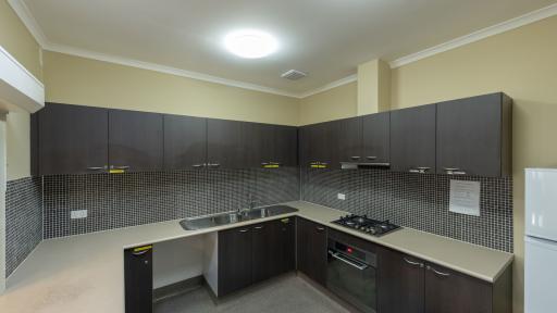Kitchen with grey benches, black cupboards and black and white chequered wall tiles. There is an oven and empty space for a dishwasher.