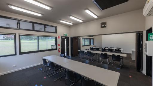 Room with three white tables and 30 black chairs. There are several wide windows on the left and a sportsground outside.