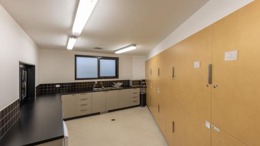 Kitchen with black benches, grey cupboards and a frosted window. There are several large brown lockers along the right.