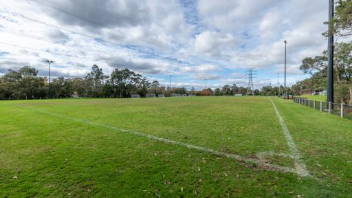 Corner view of rectangular sportsground. There are two tall light posts on the right and soccer goals and trees in the distance. Power lines are visible in a straight line above the field.