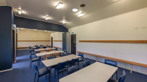 Long thin room with six white tables and 30 blue and brown chairs. There are coat racks and bench seats along the walls, and two doors to the left and right. The lights are switched on.