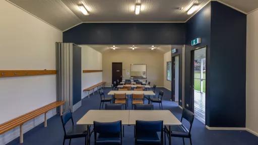 Long thin room with six white tables and 30 blue and brown chairs. There are coat racks and bench seats along the walls, two doors to right and a kitchen at the far end. The lights are switched on.