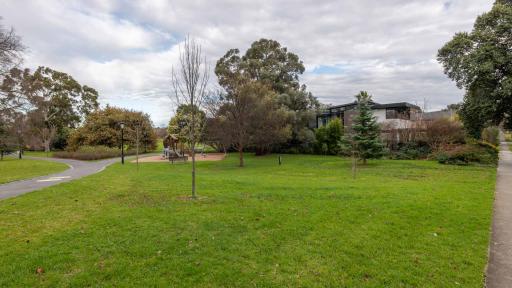 Small grass area with thick garden in far right corner and playground in far left corner. There are three small trees on the grass and walking paths on the right and left.