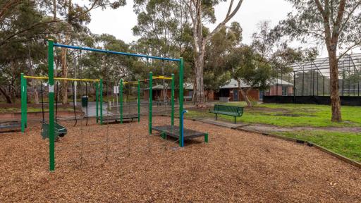 Small playground with swings and climbing features. There are tall trees to the right and on the horizon, and two house-shaped buildings and cricket nets in the background.