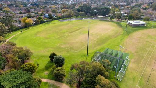 Aerial view of oval sportsground with various square line markings on its surface and four tall light posts around its edge. There is a pavilion to the top right and cricket nets to the bottom right.