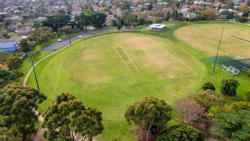 Aerial view of oval sportsground with various square line markings on its surface and light posts around its edge. There are cricket nets to the bottom right.