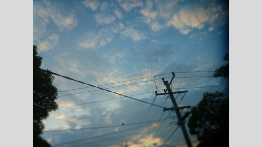 Electricity pole and cables running in several directions, against a blue sky and scattered clouds. Parts of trees are visible to the left and right.