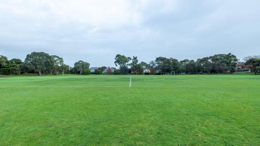 Sideline view of middle section of soccer pitch with painted white markings. There is a second adjacent soccer pitch in the background and tall trees on the horizon.