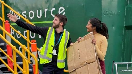 A person holding a flattened cardboard box being directed by a staff member where to put it at the recycling depot