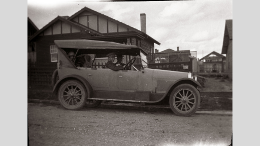 Black and white shot of early-era car with a house in the background.