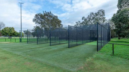 Cricket nets with 5 lanes divided by tall black fences. There are tall trees and a light post in the background.