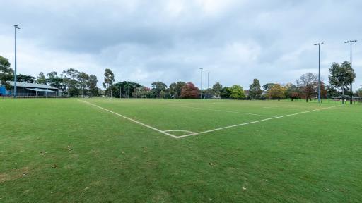 Corner view of soccer pitch with no goals at near end and AFL goal posts at far end. There is a one-storey building at the distant left and five visible light posts.