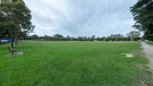 Large grass field with dirt walking track to the right and a long row of medium trees and park benches to the left. There are soccer goals in the distance.