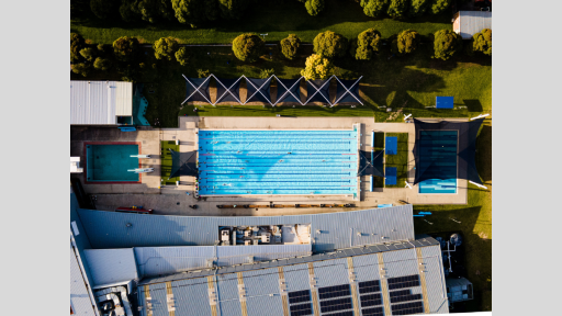 Aerial shot of swimming pool with black lap lines. There is a building at the bottom and lush trees at the top.