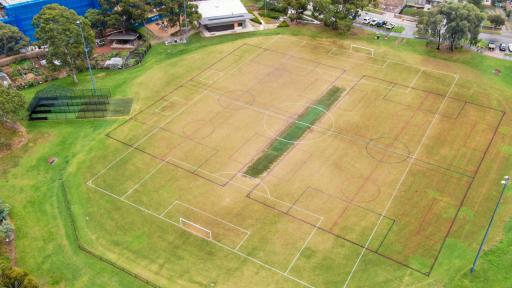 Aerial view of sports field with white markings for soccer and black markings for another sport. There are cricket practice nets to the left, a pavilion at the top and a road to the top right.