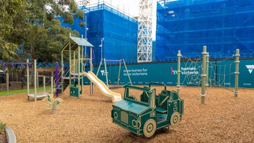 Playground with small climbing feature, slide, swings, toy car and rope feature. There is a fenced-off construction area and crane in the background.