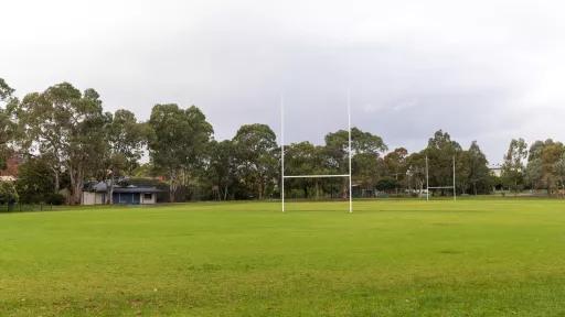Sports field with rugby goal posts at each end and grass area in the foreground. There is a single-storey dark blue and cream white building to the far left and tall trees in the background.