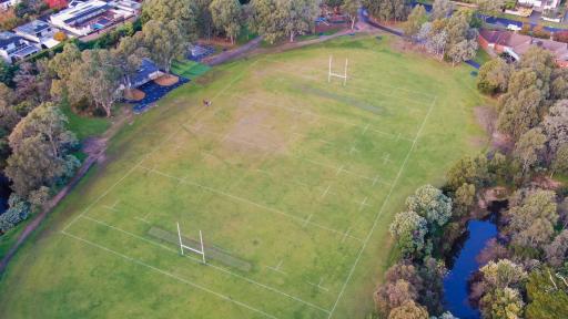 Aerial view of playground to the south and sportsground with rugby goal posts to the north. There are walking paths on each side of the playground and thick mature trees surround the area.