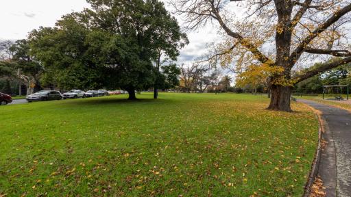 Grass area with fallen leaves and several scattered large trees. There is a curved walking path and part of a playground to the right and a line of parked cars to the left.