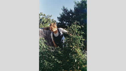 Woman in dark blue overalls and white t-shirt looking at thick garden trees.