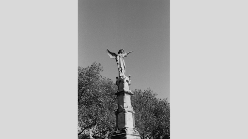 Black and white shot of tall concrete statue with winged woman on top. There are trees in the background.