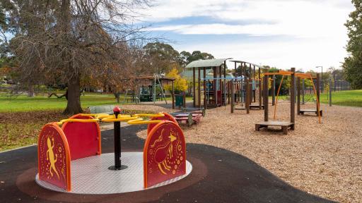 Playground with round spinning feature in foreground and climbing feature in the background. There is a large bare tree to the left.