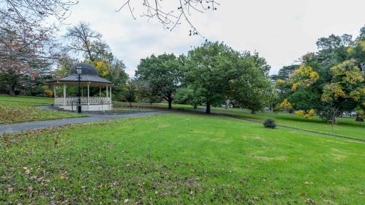 Large grass area with a left-to-right slope and a rotunda and walking path to the left. There are medium-sized trees in the background and fallen leaves in the foreground.