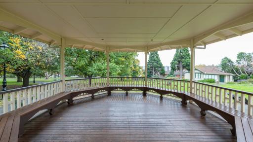 Inside view of rotunda in grass area. The columns and bench seats are dark red and the roof and balustrades are white. There are large trees in the background.