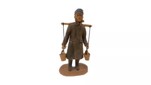 Clay sculpture of a man wearing a long dark grey coat, with dark grey boots and cap. Work uniform including a white shirt, dark grey vest, pants, boots and cap. A long stick is balanced horizontally on his back with two pieces of twine connected to clay buckets tied at both ends. The man’s hands are loosely griped on both pieces of twine, just above the dangling buckets.