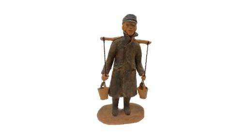 Clay sculpture of a man wearing a long dark grey coat, with dark grey boots and cap. Work uniform including a white shirt, dark grey vest, pants, boots and cap. A long stick is balanced horizontally on his back with two pieces of twine connected to clay buckets tied at both ends. The man’s hands are loosely griped on both pieces of twine, just above the dangling buckets.