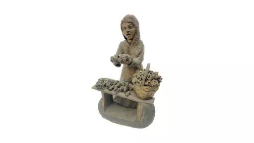 Clay sculpture of a woman painted entirely in dark brown glaze. She is wearing a long dress and standing behind a bench. On the bench is a basket filled with flowers and the rest of the bench is also covered with flowers. The woman’s mouth is open as if she id pleading, and both hands stretched out in front, holding more flowers.